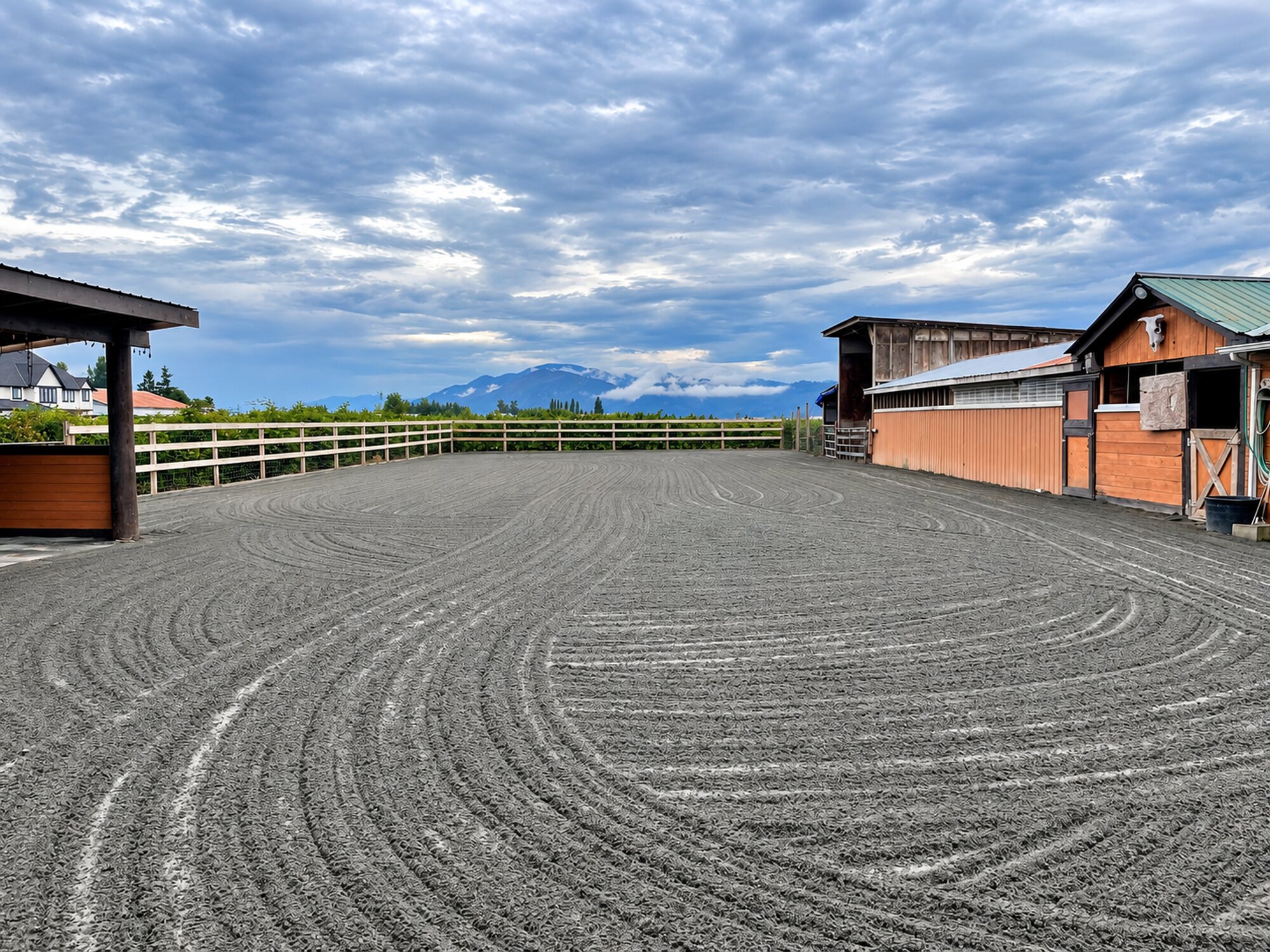 Freshly graded riding arena with an even prepared surface, perimeter fencing, barns, and mountains beyond